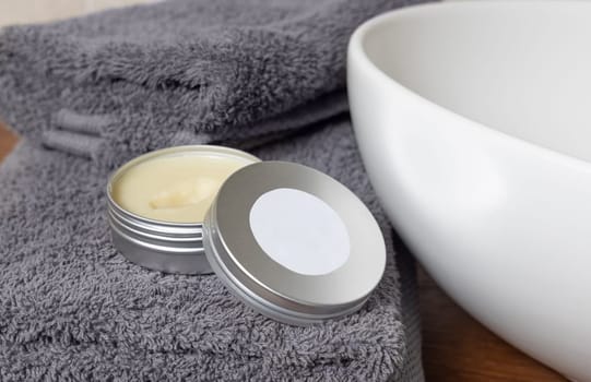 White opened body butter jar with label near dark grey folded towel and vessel basin on brown wooden countertop in bathroom, close up, cosmetic mockup for brand packaging