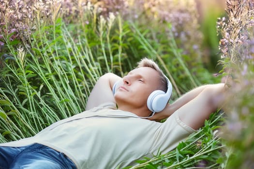 Man relaxing lying in a field among flowers and listening to music in headphones with eyes closed.