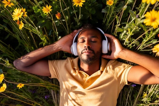 Relaxed black man relaxing lying on lawn among flowers listening to music in headphones with eyes closed, enjoying summer and nature
