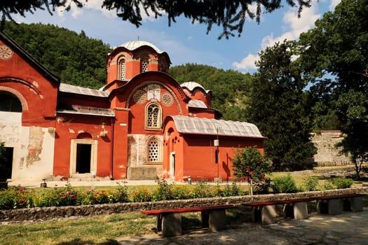 View onto the elaborate front facade and roof, domes, cupolas of the bright red Church Complex inside the courtyard, garden of the Patriarchate of Pec Monastery with a bench in the foreground, outside of Peja, Kosovo 2022
