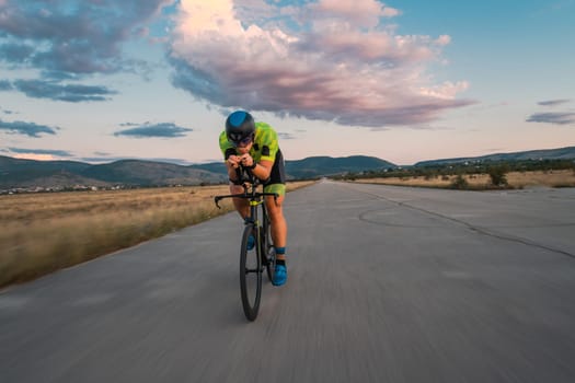 Triathlete riding his bicycle during sunset, preparing for a marathon. The warm colors of the sky provide a beautiful backdrop for his determined and focused effort