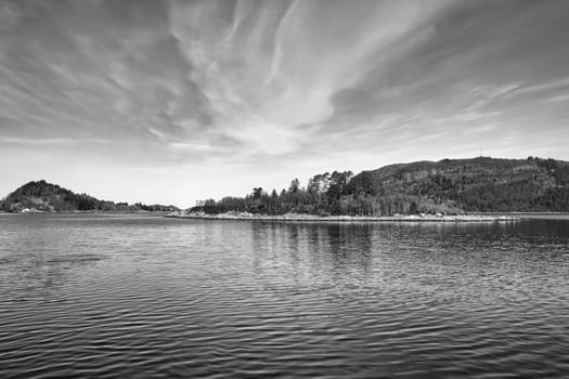 Island with rocks and trees in the fjord in front of the open sea in Norway in black and white. The water glistens in the sun. Landscape from the north