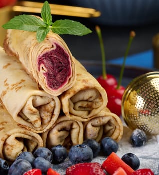 Crepes stacked in a pile, one of which is cut and filled with berry filling. The plate is also decorated with blueberries, strawberries, and cherries, close up