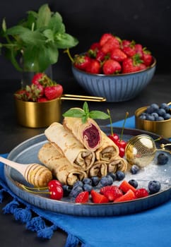 Crepes stacked in a pile, one of which is cut and filled with berry filling. The plate is also decorated with blueberries, strawberries, and cherries, a honey dipper with honey