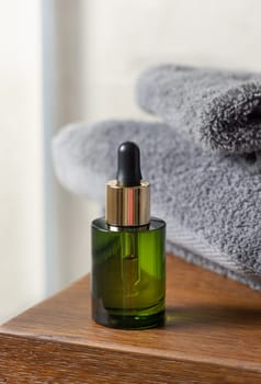 Green dropper bottle near grey folded towel on vessel basin on brown wooden countertop in bathroom, close up, cosmetic mockup for brand packaging