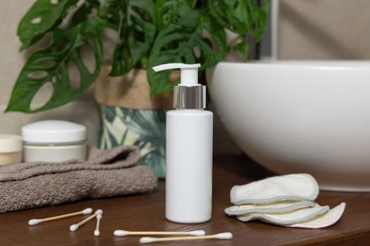 White pump bottle near basin and green monstera plant on wooden countertop in bathroom close up, cosmetic mockup. Personal care products for brand packaging