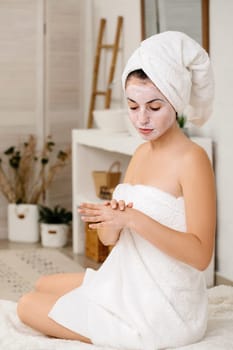 young woman with towel is applying mask on her face at home. Skin care.