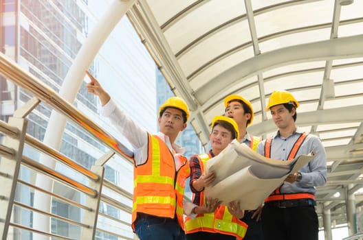 Team of engineers and architects four male wearing safety yellow helmet and reflective clothing standing in front of modern building. Project engineering manager in white shirt pointing finger to construction building for successful project. Construction and engineering concept.