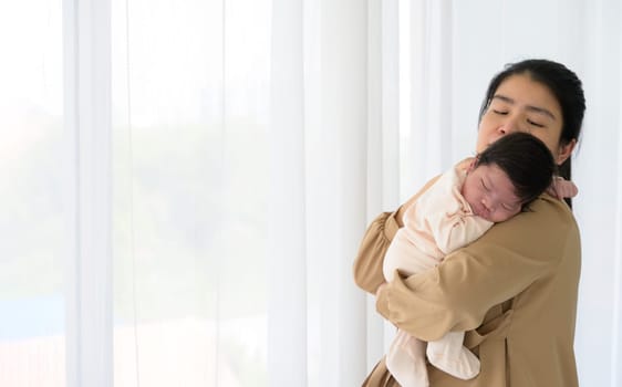 Newborn baby in tender embrace of mother at white curtain window. Adorable infant girl 1 month in jumpsuit sleeping on mom shoulder at home. Care, love and mothers day