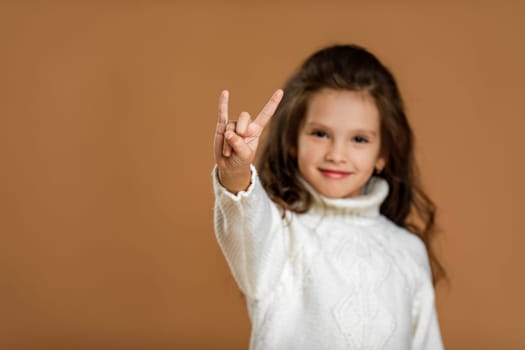 Cute smiling little child girl in white sweater making Rock gesture on beige background. focus on hand