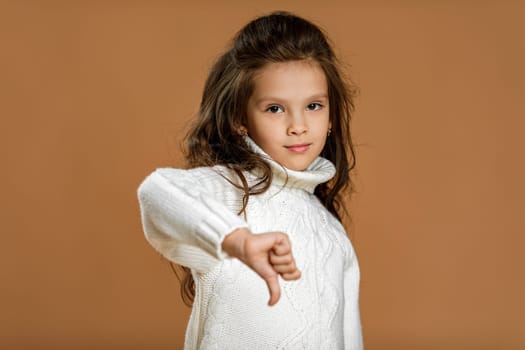 Cute little child girl in white sweater showing the thumbs down gesture on beige background.