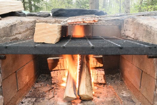 A close-up view of a brick outdoor fireplace with a metal grate, wood logs burning underneath, and a grilling basket on top.
