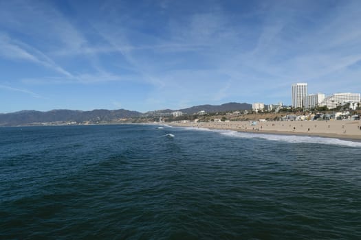 A scenic view of a beach with clear blue water and a cityscape in the background. The sky is clear with a few wispy clouds. People are walking along the sandy shore.
