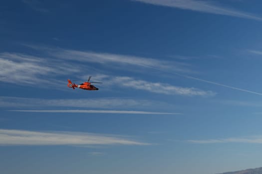 A bright orange helicopter flying in a clear blue sky with wispy clouds.