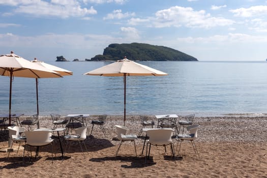 Open empty cafe on the beach with tables, chairs and umbrellas set up on the sand. Calm sea and island in the background.preparing for the season. Copy space.