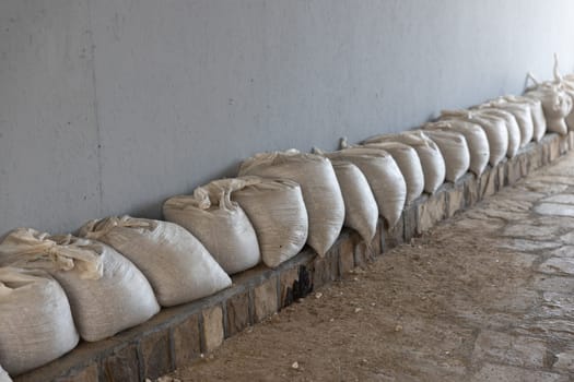 A row of white sandbags lined up against a wall on a stone floor for flood protection. The sandbags are neatly arranged against a plain wall. Copy space.