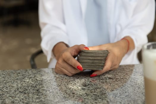 Woman with red nails and white blouse holding a stack of tarot cards at a granite table in a cafe. Copy space.