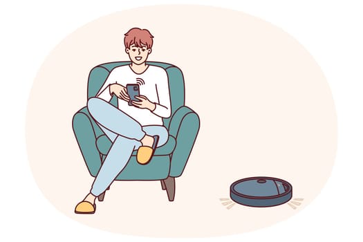 Robotic vacuum cleaner guided using mobile phone cleans floor in apartment near man sitting in chair. Robot vacuum cleaner with wi-fi chip is controlled via IOT and integrated into smart home system