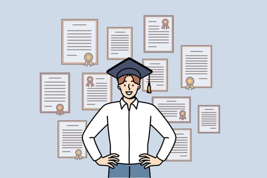 Man graduate stands in academic hat near wall with diplomas from advanced business courses. Guy is graduate and proud of good education and availability of documents confirming qualifications