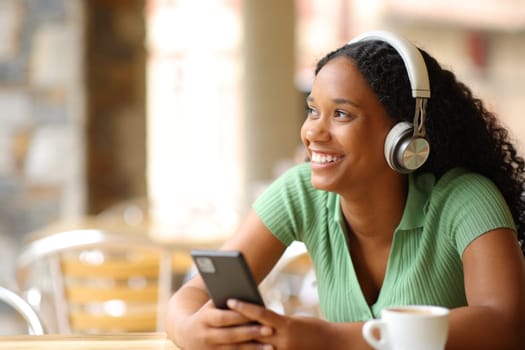Happy black woman listening audio holding phone looking at side in a bar terrace