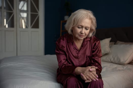 Unhappy sad elderly woman wearing pajamas sitting on bed feeling loneliness having psychological and mental troubles