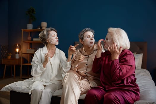 Senior women applying anti-aging cotton facial mask having fun together. Positive relaxed female best friends feeling cheerful and happy spending time on weekends