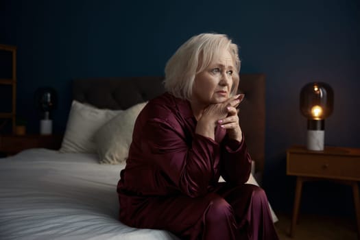 Sad elderly woman wearing pajamas sitting on bed feeling loneliness having psychological and mental troubles
