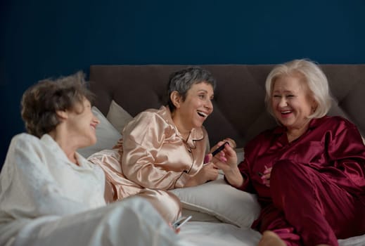 Attractive retired female friends applying nail polish while having playful pajamas party time at home. Friendship, positivity and nice communication on retirement