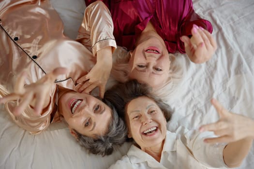 Top view on three positive smiling elderly woman friends wearing pajamas lying in bed. Hen-party on retirement