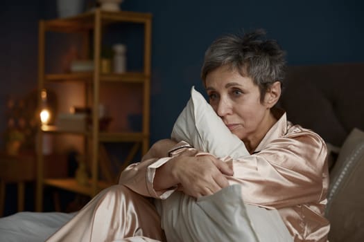Depressed elderly woman suffering from psychological problem feeling frustration and melancholy hugging pillow sitting on bed