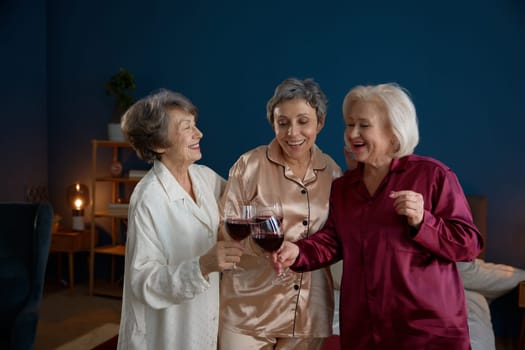Female senior friends in pajamas drinking wine enjoying fun retirement time together. Healthy attractive mature women speaking gossiping meeting on slumber party at home