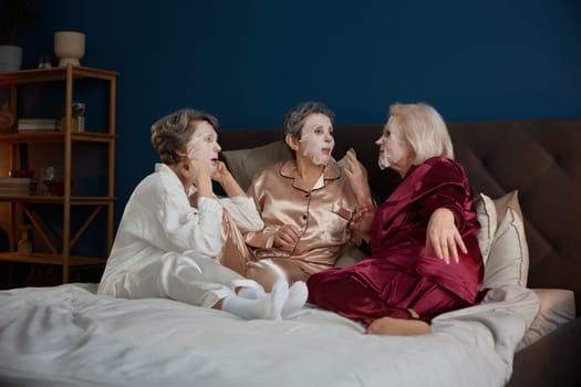 Happy senior female friends with cotton mask on face having nice conversation. Aged lady applying anti-aging treatment cosmetics enjoying wonderful time spent together during pajama party at home