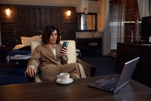 Young adult businesswoman using phone and laptop for work in luxury hotel room. Elegant female entrepreneur taking coffee break during busy job evening during business travel