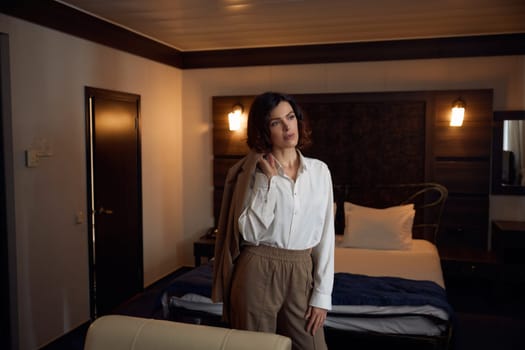 Portrait of thoughtful elegant businesswoman holding suit jacket on shoulder looking confidently into window in hotel bedchamber. Business voyage concept
