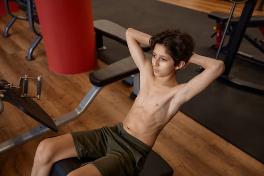 Weak skinny preteen boy pumping up his abs doing fitness exercise to make body strong, sportive and healthy. Workout motivation and gym training for children