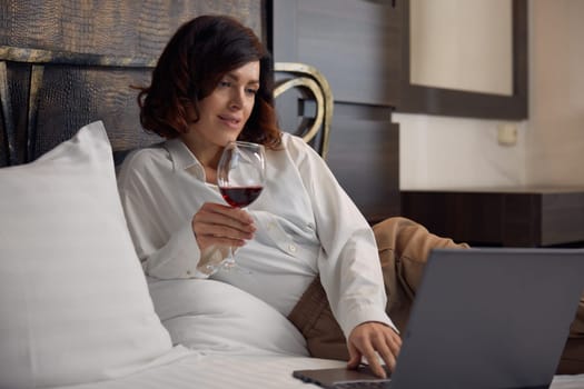 Relaxed businesswoman lying in bed drinking wine and browsing internet on laptop computer enjoying evening in hotel room