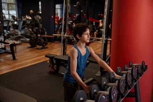 Pensive overwhelmed preteen boy at dumbbells having choice making decision about weights for training at gym