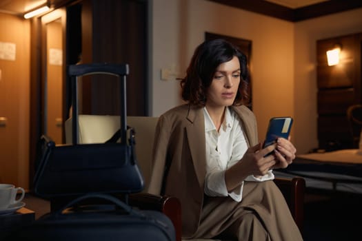 Young businesswoman typing message on smartphone booking return ticket while sitting with luggage bag and suitcase in hotel room. Finish of business trip concept
