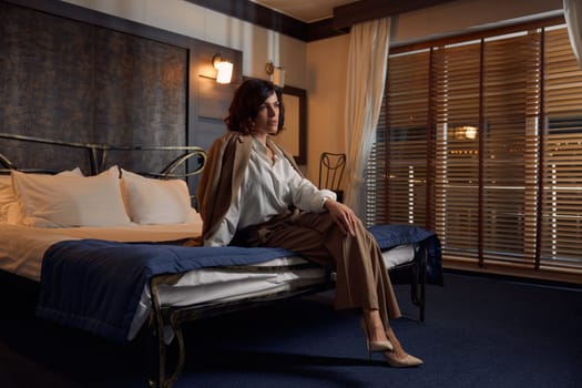 Young confident businesswoman sitting on bed in hotel room. Female executive dreaming and relaxing after business conference or corporate partners meeting