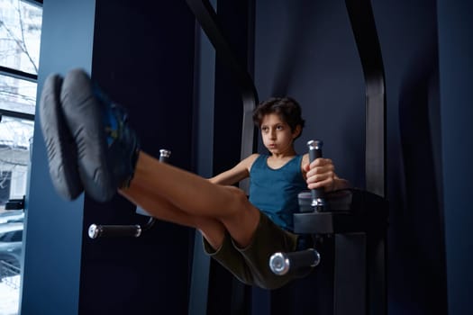 Sporty preteen boy training abs demonstrating strength and power. Muscular schoolboy showing endurance and effort during workout at gym