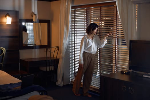 Worried businesswoman looking through blinds while standing in hotel room. Hostel guest feeling nervous before business meeting or company partners conference