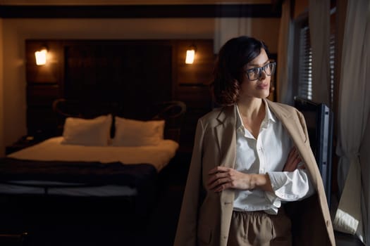 Successful joyful businesswoman with suit jacket on shoulders looking dreamy at window in hotel room. Attractive lady planning next day during business trip adventure