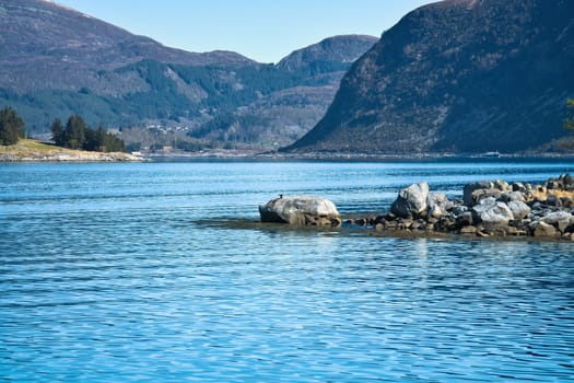 Fjord with mountains on the horizon. Stones reach into the water. The water glistens in the Norwegian sun. Landscape photo from the north