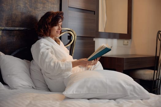 Businesswoman wearing bathrobe reading book lying in bed surrounded with soft pillow while rest in hotel room