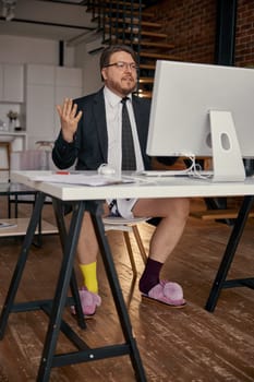 Serious businessman working online on computer wearing formal suit and funny home slippers sitting at table. Business meeting, briefing or interview, remote job or freelancing