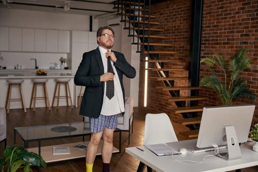 Funny businessman wearing formalwear and different socks knotting necktie preparing for online work standing at home office. Freelance occupation, remote job, business in cyberspace