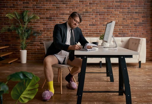 Nerd funny businessman working online on computer wearing formal suit and funny home slippers sitting at table. Business meeting, briefing or interview, remote job or freelancing