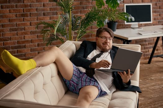 Surprised businessman grimacing looking at laptop screen working online sitting on sofa at home living-room. Confused salesman entrepreneur reading e-mail news using computer