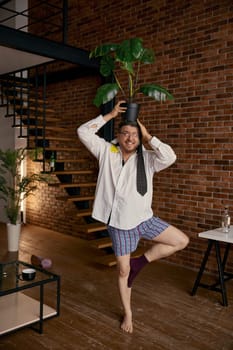 Crazy drunk businessman or corporate online worker with flowerpot on head fooling around standing on one leg at home living room. Remote employee drunkard