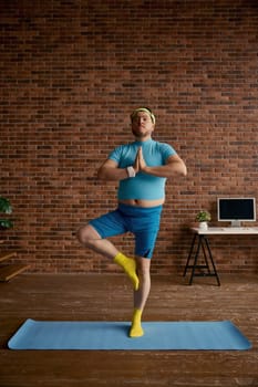 Relaxed fat sportsman meditating standing on mat at home. Overweight man calming down practicing yoga position. Sports activity and weight loss